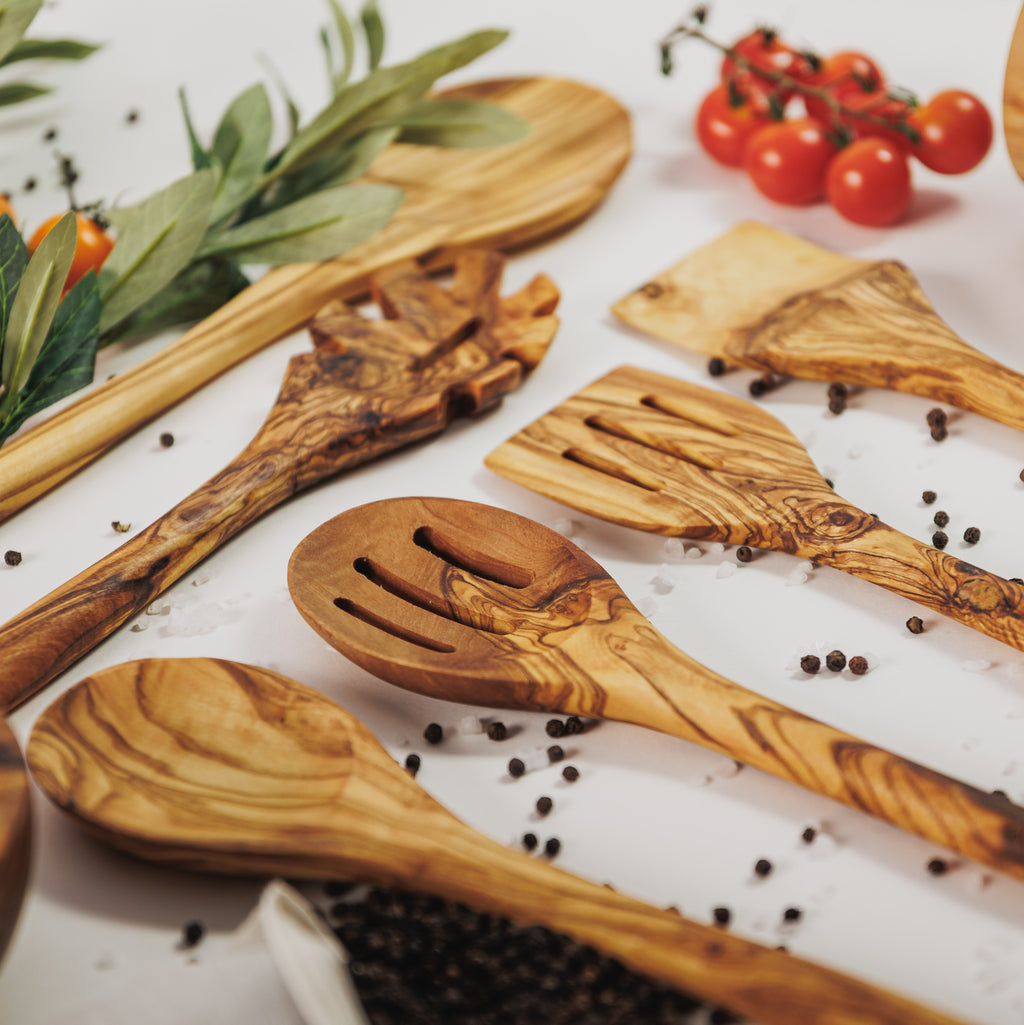 Assortment of olive wood cooking utensils including spoons, spatulas, and slotted servers, styled with cherry tomatoes, peppercorns, and olive branches on a white surface