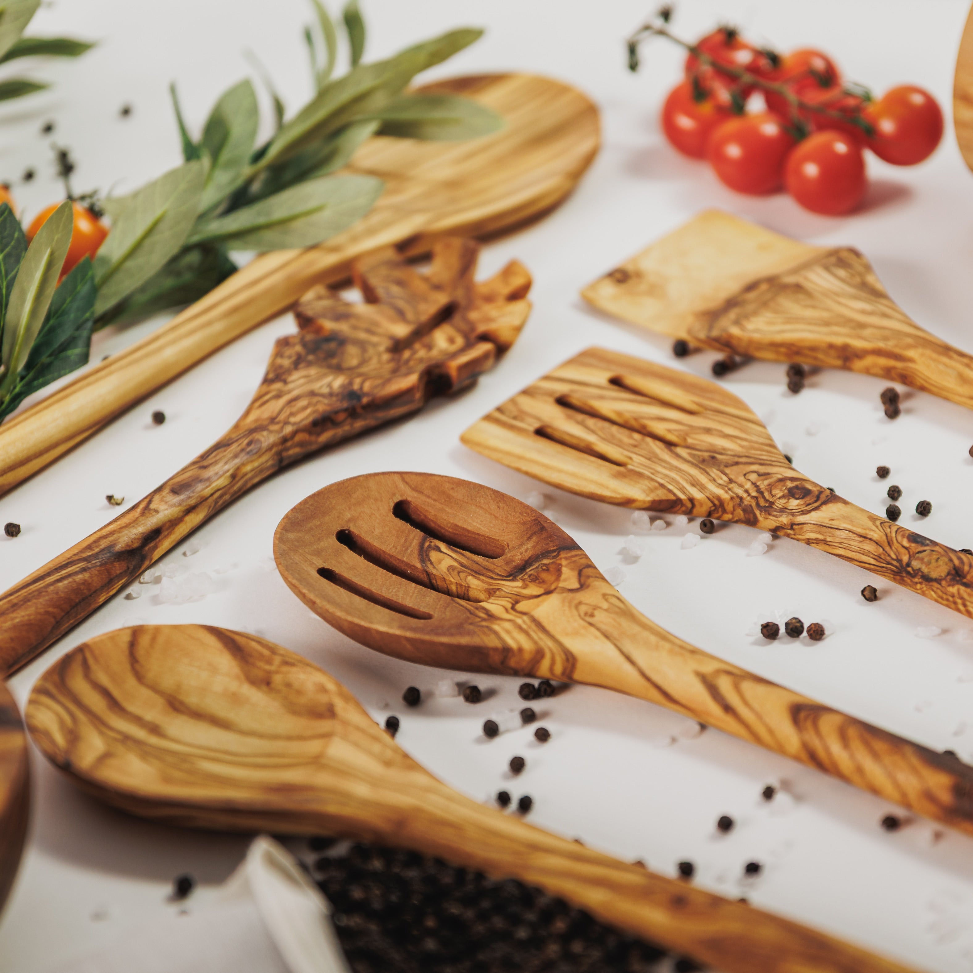 Assortment of olive wood cooking utensils including spoons, spatulas, and slotted servers, styled with cherry tomatoes, peppercorns, and olive branches on a white surface