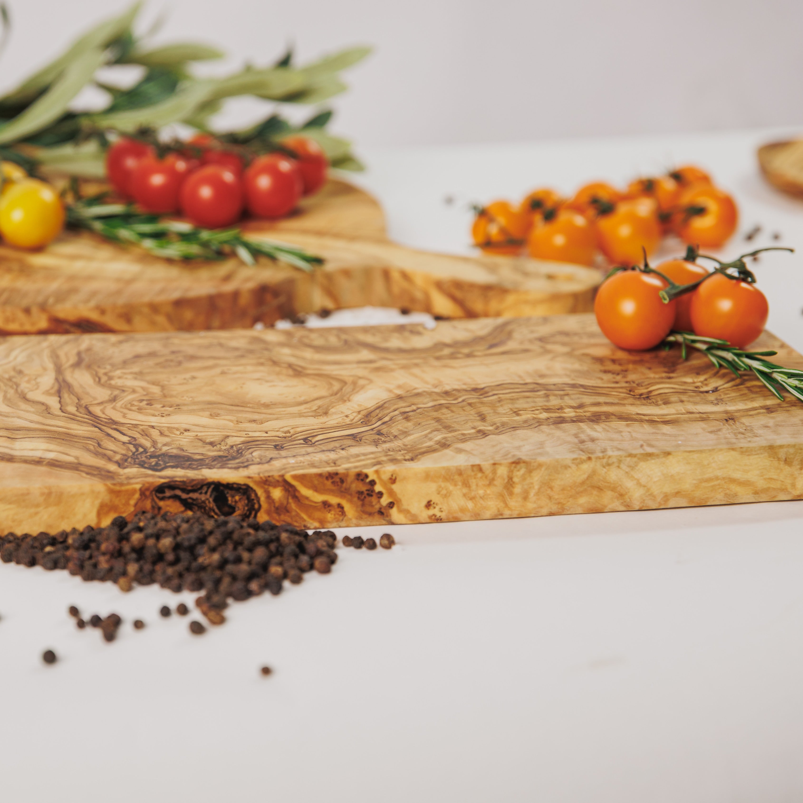 Close-up of two olive wood cutting boards showing the natural grain detail, styled with cherry tomatoes, rosemary, and peppercorns on a white surface