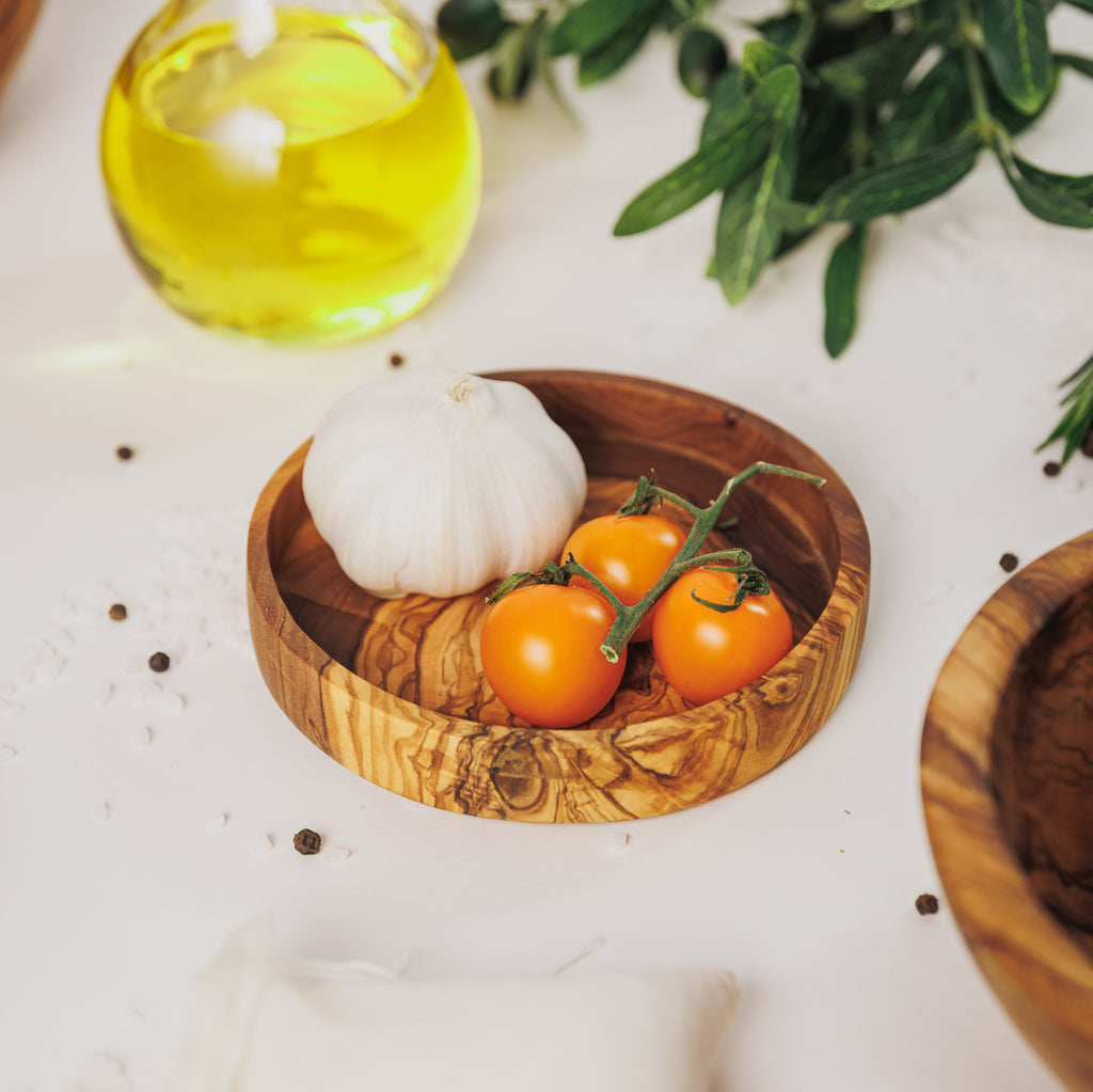 Olive wood dipping dish holding a garlic bulb and yellow cherry tomatoes, styled with a bottle of olive oil and olive branches on a white surface