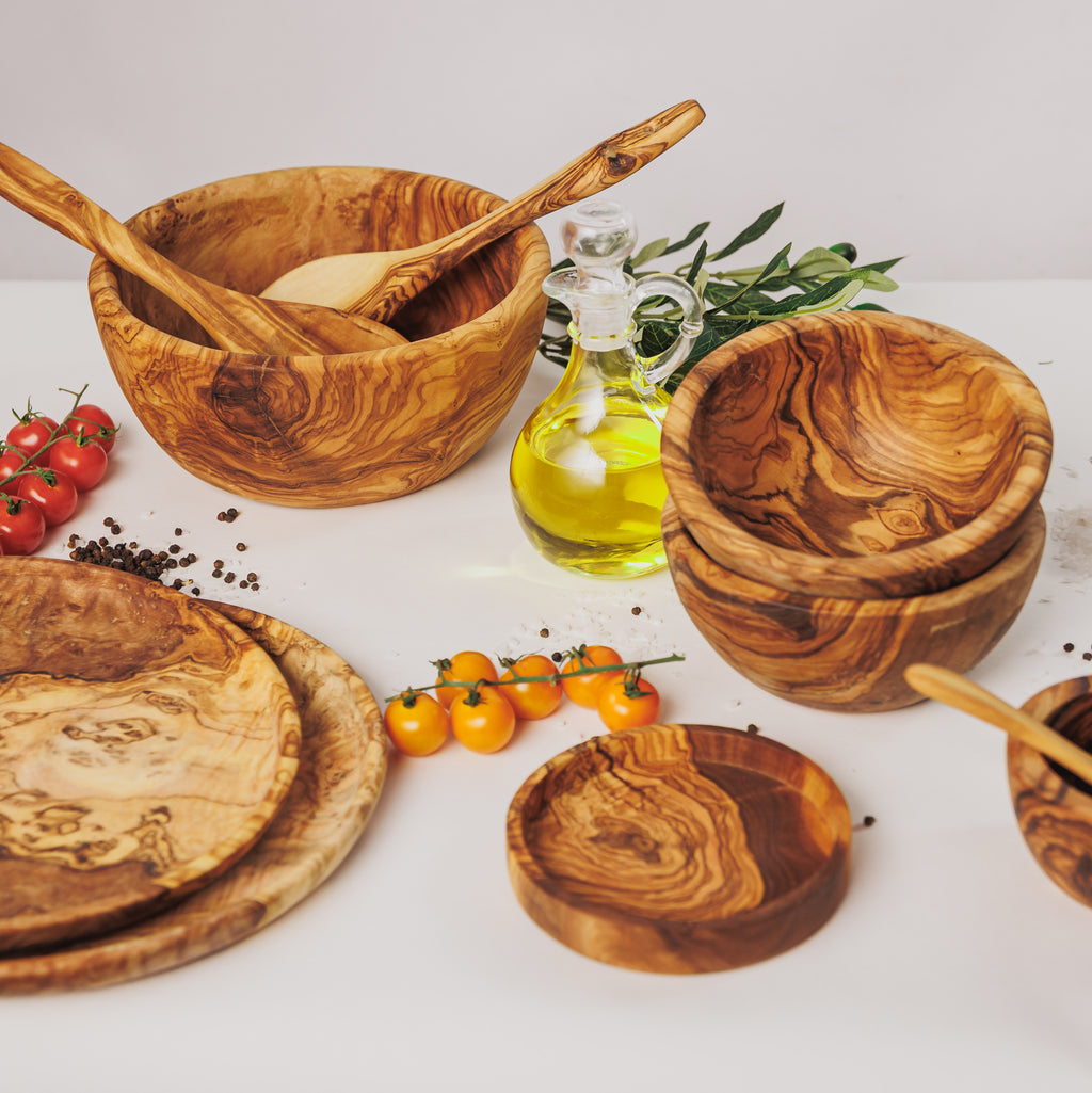 Collection of olive wood kitchenware including salad bowl, serving utensils, small bowls, plates, and cutting boards, styled with cherry tomatoes, peppercorns, and a bottle of olive oil on a white surface