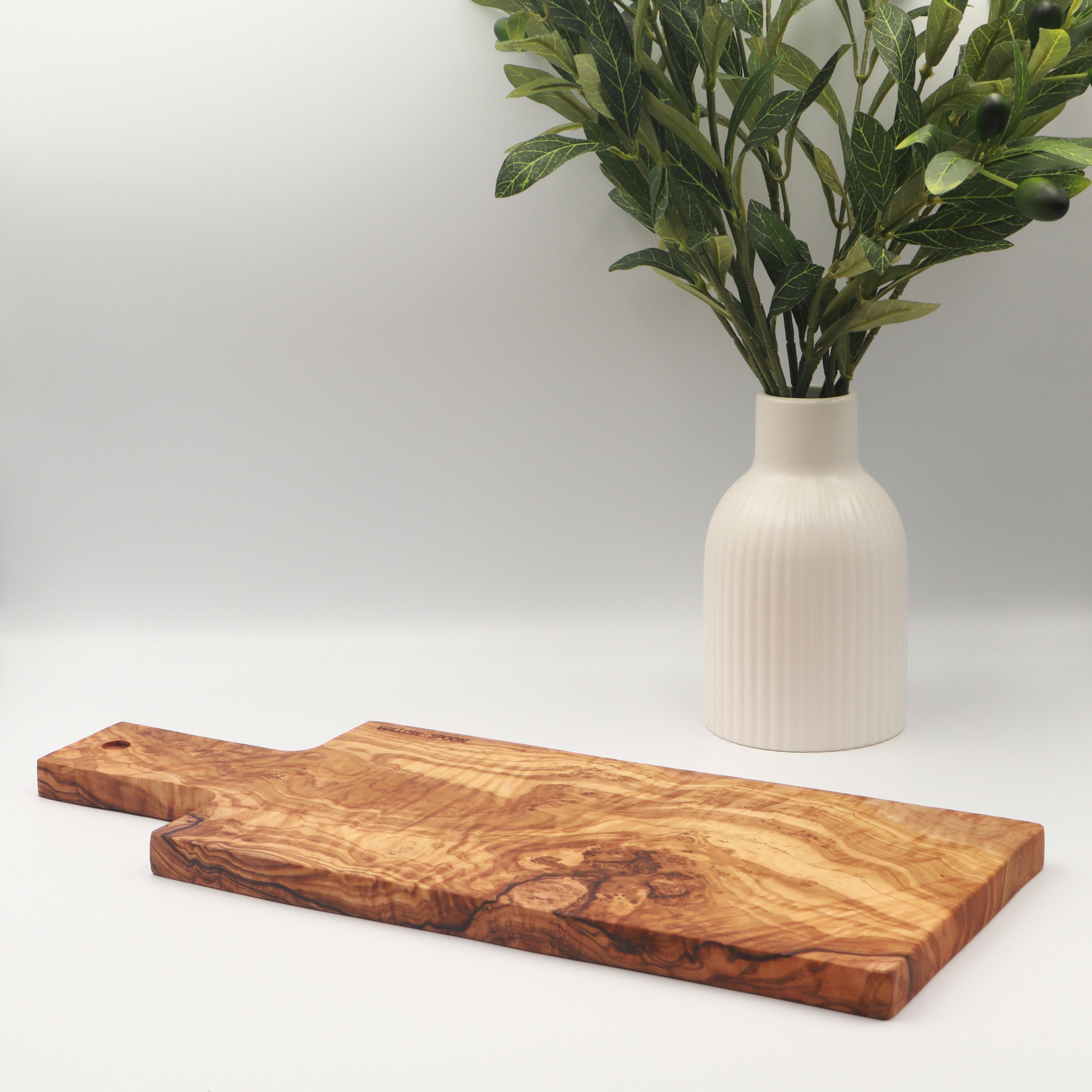 Handcrafted Tunisian olive wood rectangular cutting and serving board with handle, styled on a white counter beside a vase of olive branches.