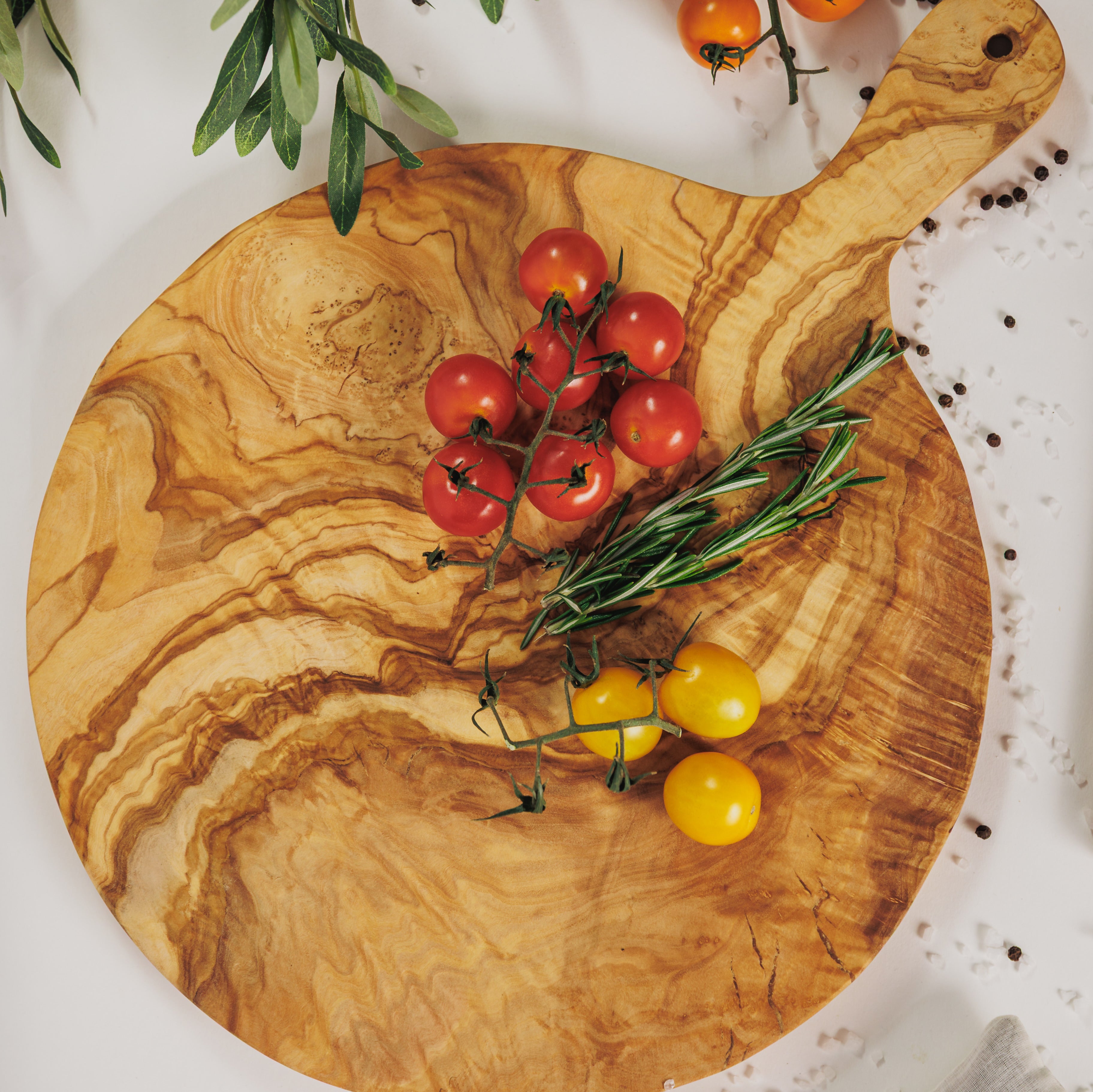 Round olive wood cutting board with handle, styled with cherry tomatoes and rosemary on a white surface