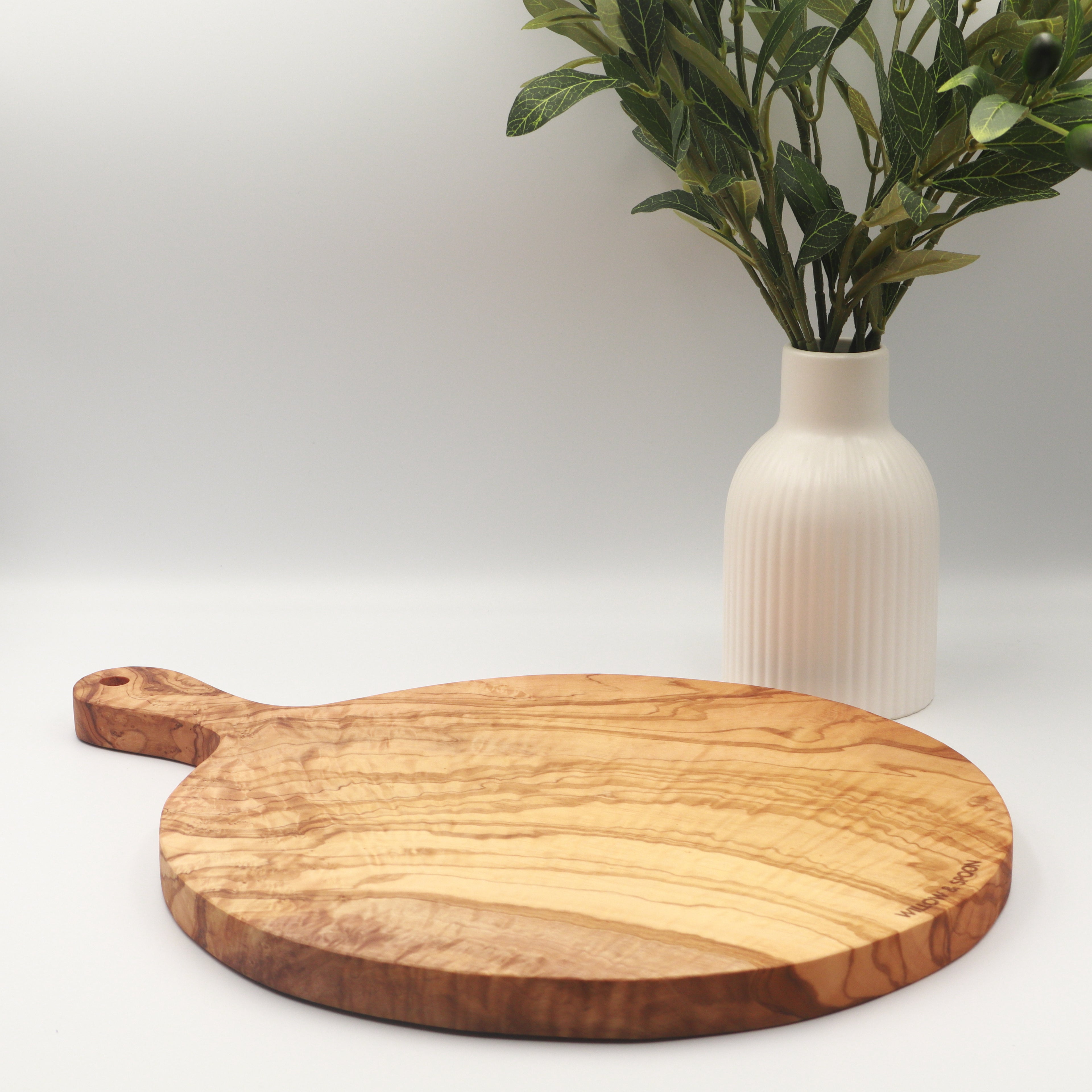 Handcrafted Tunisian olive wood round cutting board with handle, styled on a white counter beside a vase of olive branches.