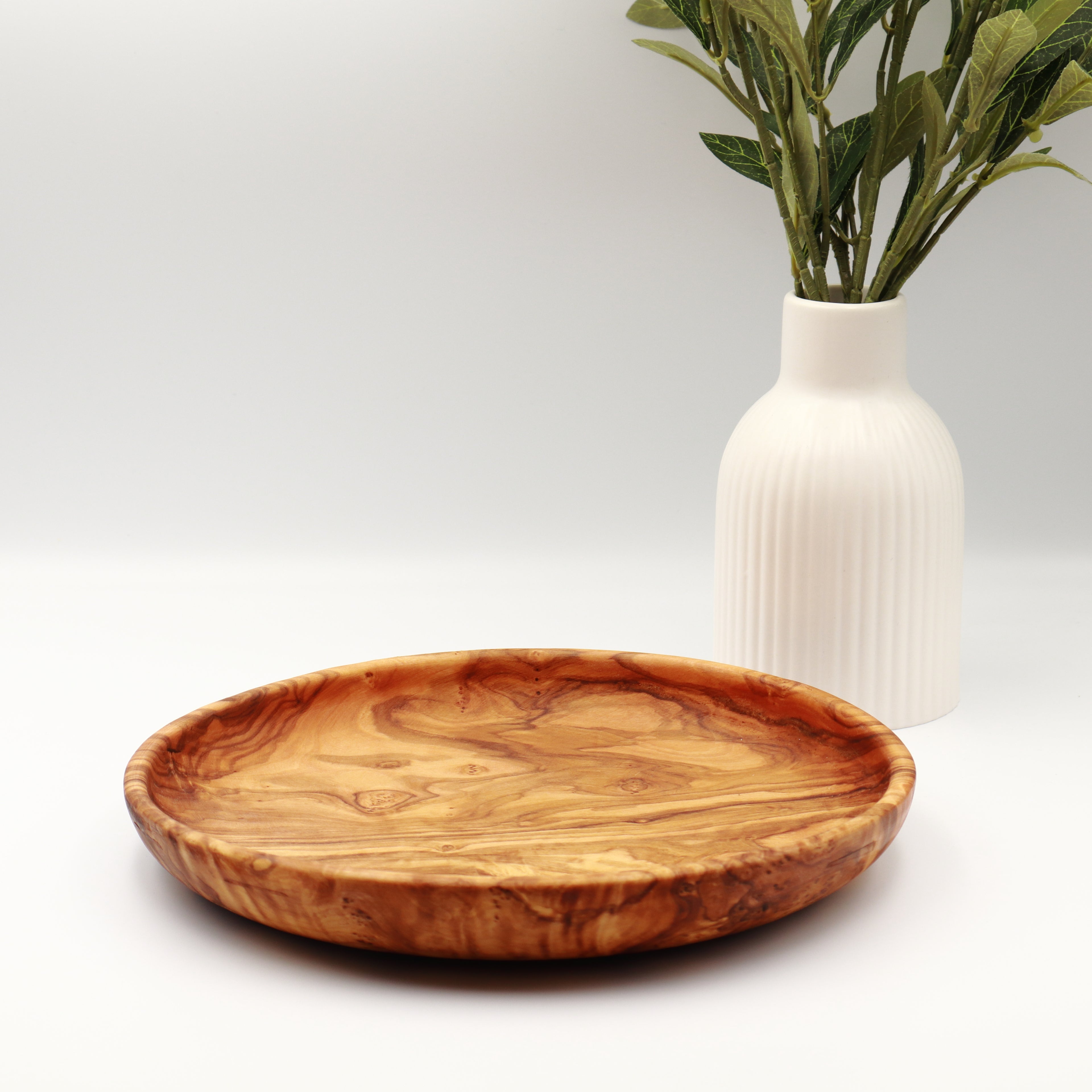 Handcrafted Tunisian olive wood round plate, styled on a white counter beside a vase of olive branches.