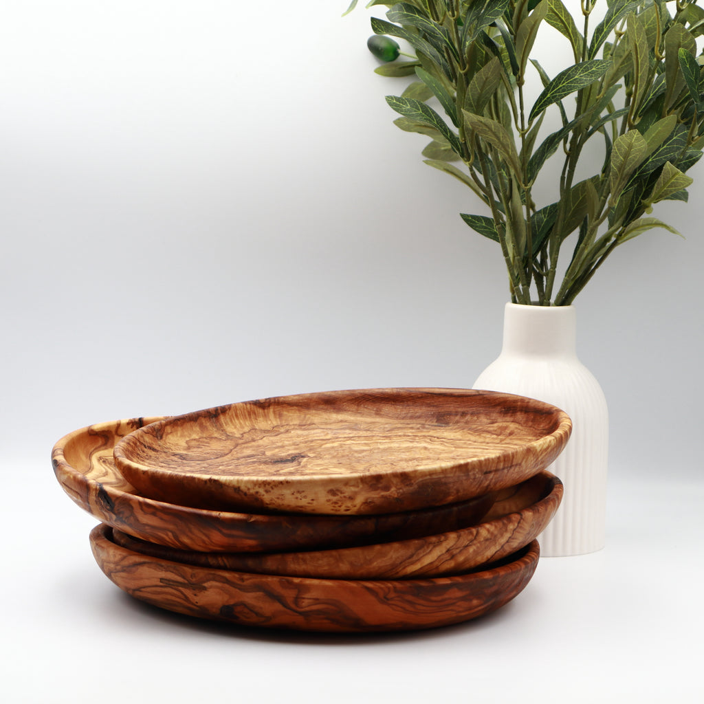 Four stacked handcrafted Tunisian olive wood round plates, styled on a white counter beside a vase of olive branches.