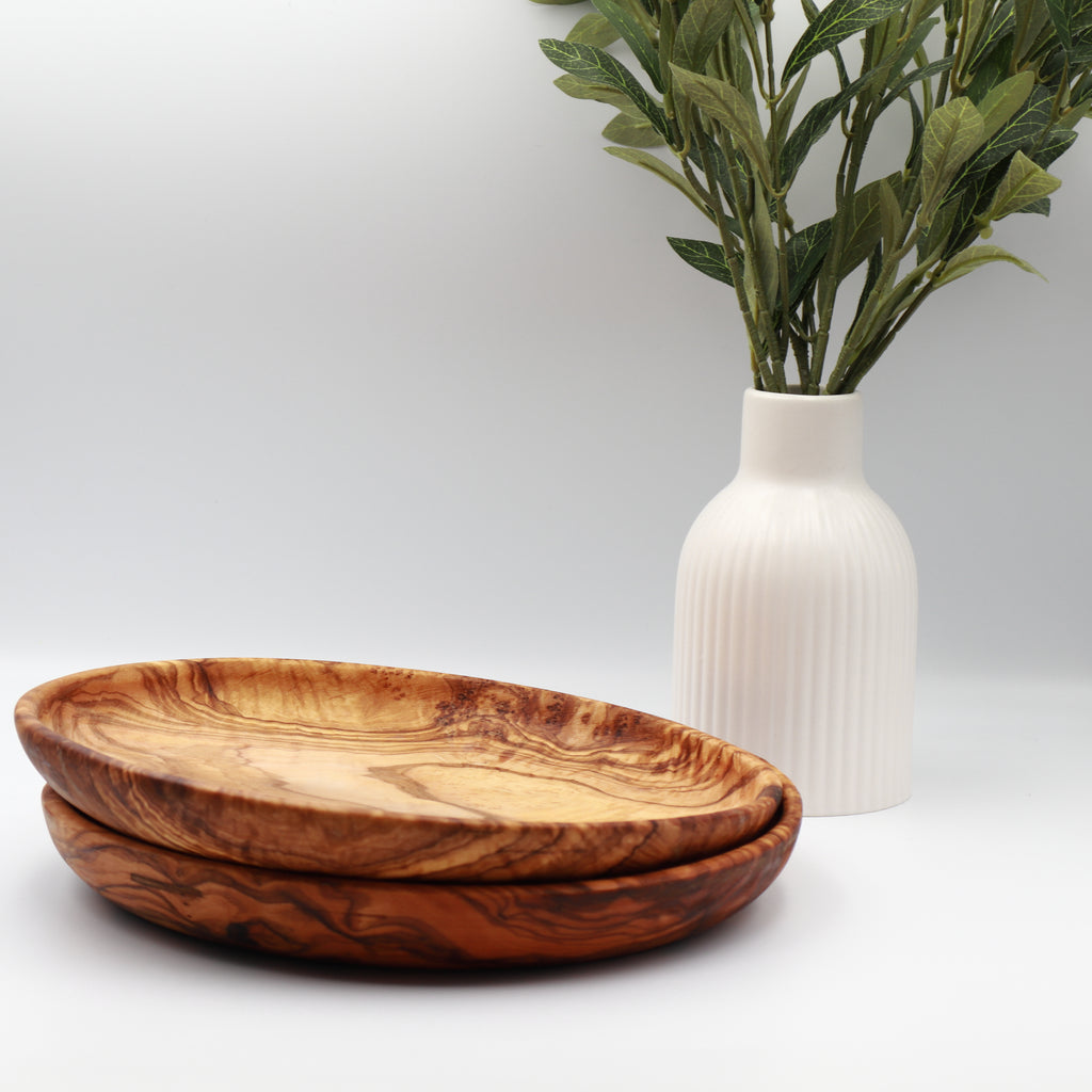 Two stacked handcrafted Tunisian olive wood round plates, styled on a white counter beside a vase of olive branches.