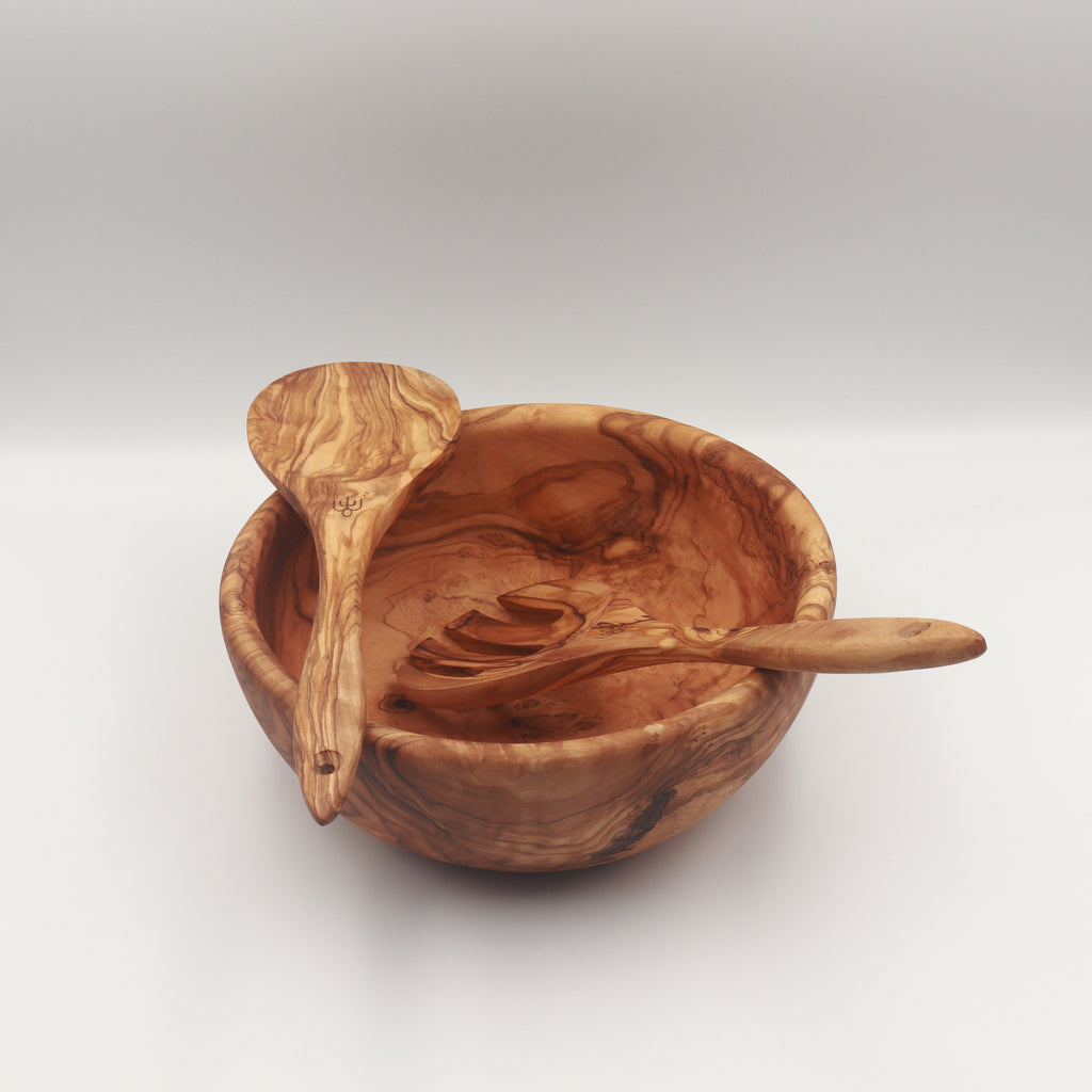 Angled view of a handcrafted Tunisian olive wood salad bowl with two salad servers resting inside on a white background.