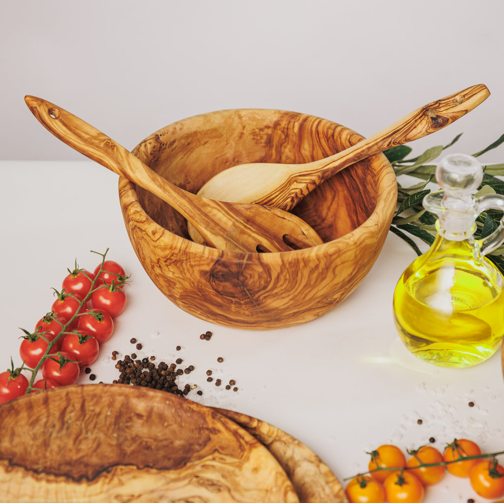 Olive wood salad bowl with serving utensils resting inside, styled with cherry tomatoes, peppercorns, and a bottle of olive oil on a white surface
