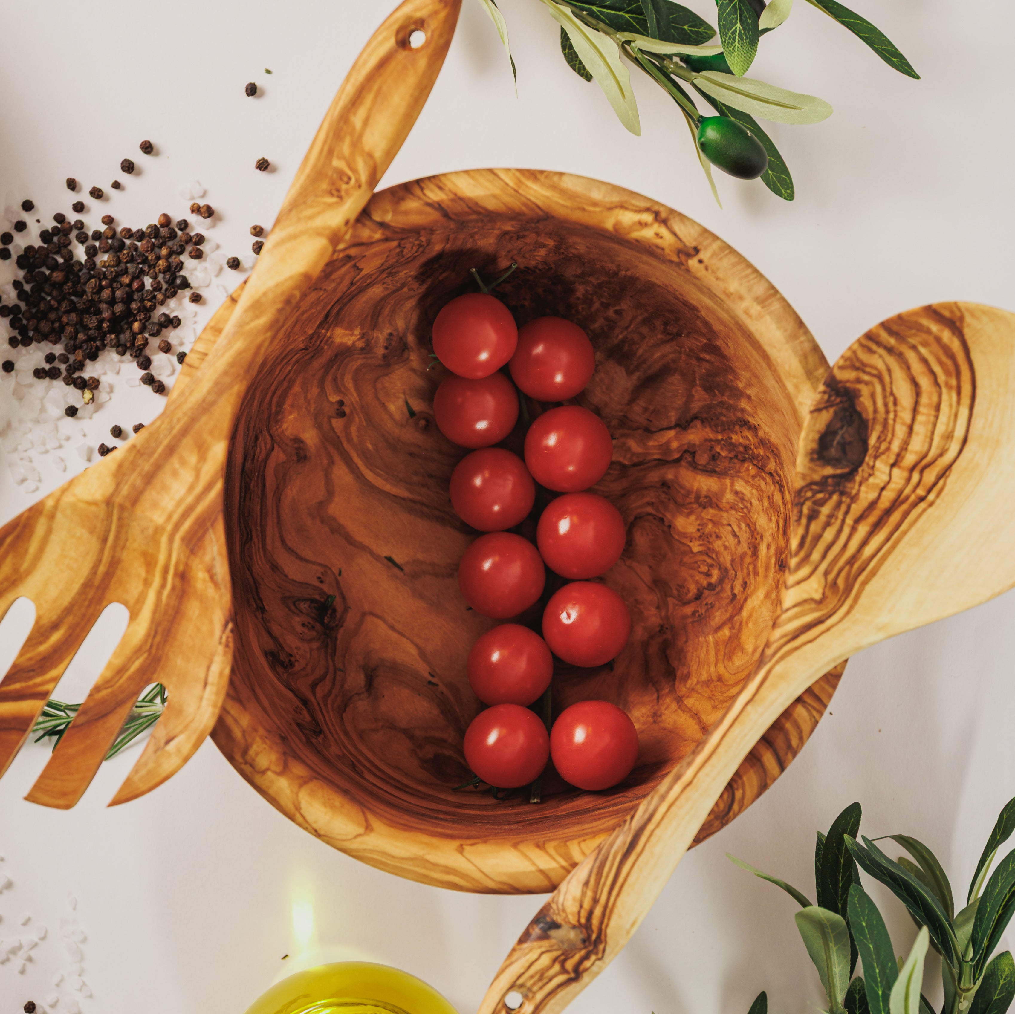 Olive wood salad bowl filled with cherry tomatoes, flanked by matching serving utensils, peppercorns, and olive branches on a white surface