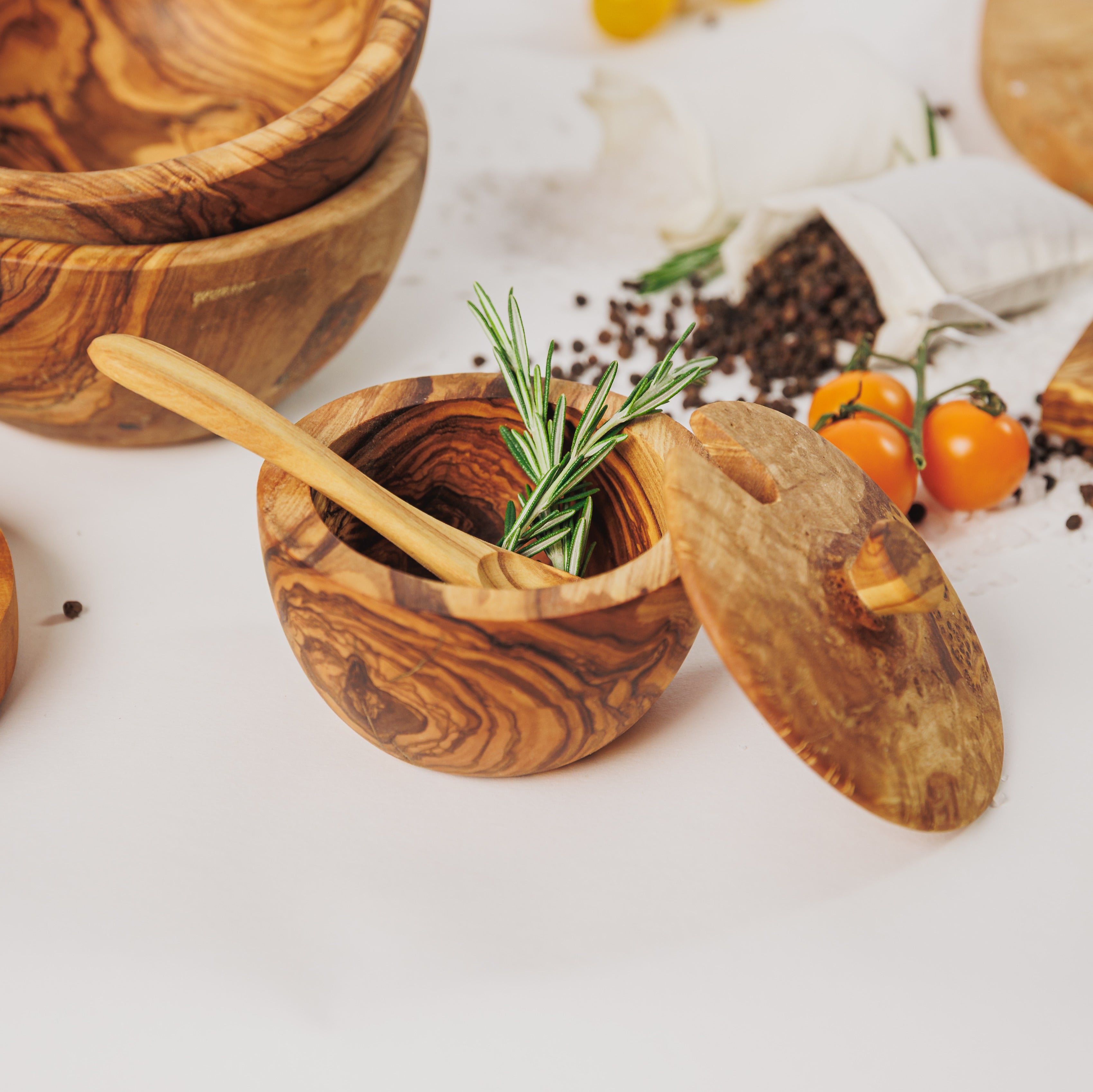 Olive wood salt cellar with a small spoon and swivel lid, styled with rosemary, peppercorns, and cherry tomatoes on a white surface