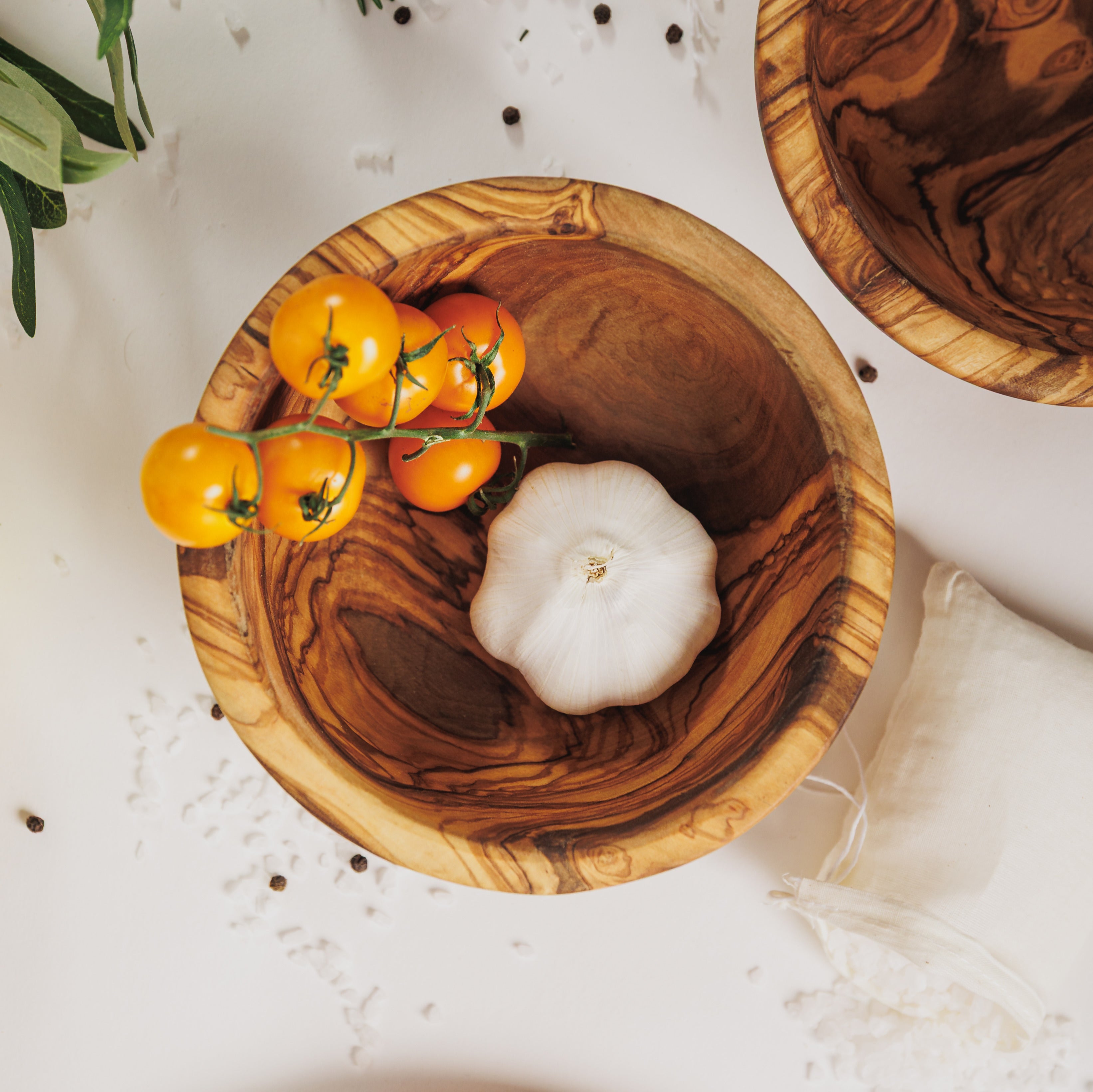 Olive wood serving bowl holding a garlic bulb and yellow cherry tomatoes, with a matching bowl and olive branches on a white surface