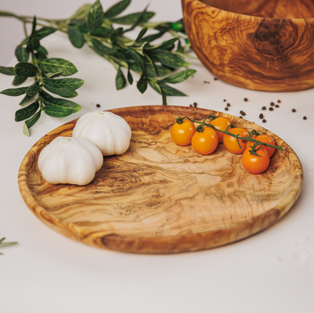 Olive wood serving plate holding garlic bulbs and yellow cherry tomatoes, with a matching bowl and olive branches in the background on a white surface