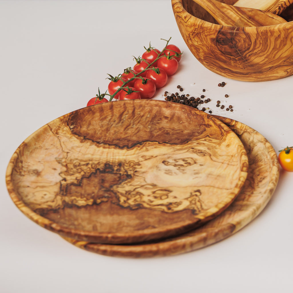 Stacked olive wood serving plates, styled with cherry tomatoes, peppercorns, and a matching salad bowl on a white surface