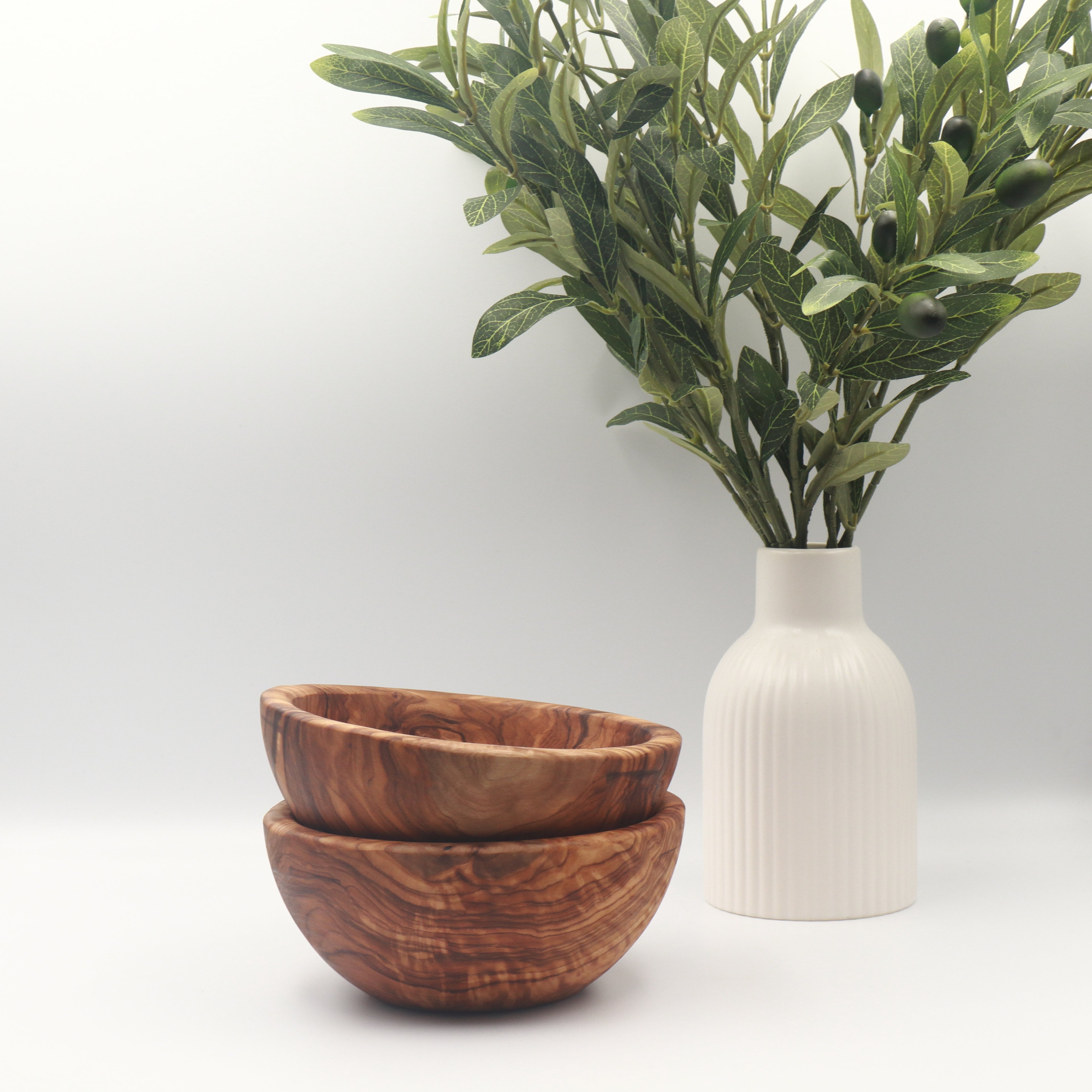 Two stacked handcrafted Tunisian olive wood small bowls, styled on a white counter beside a vase of olive branches.