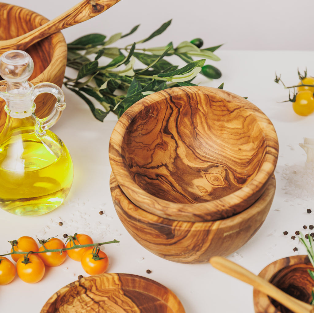Stacked olive wood small bowls, styled with a bottle of olive oil, yellow cherry tomatoes, peppercorns, and olive branches on a white surface