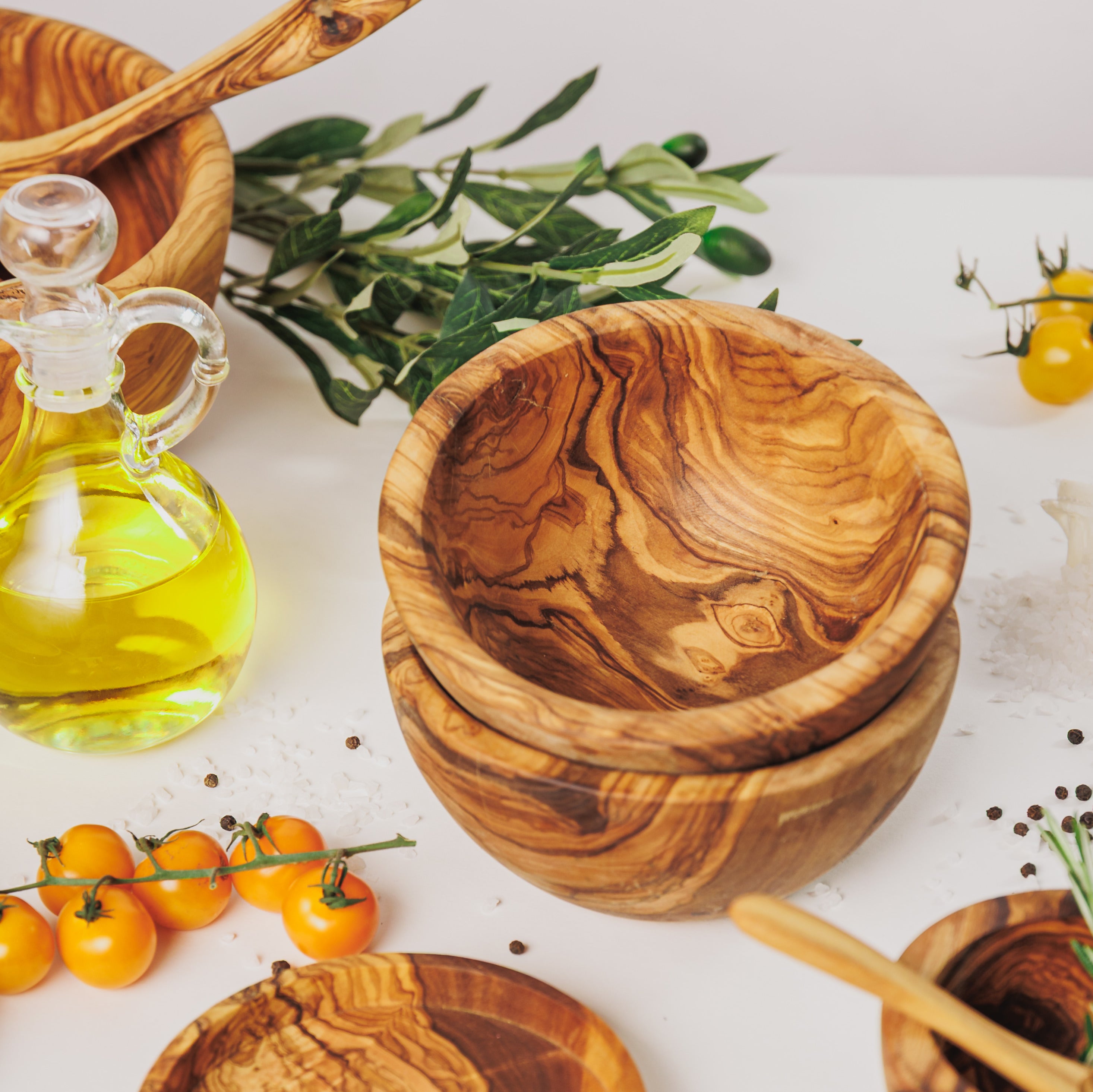Stacked olive wood small bowls, styled with a bottle of olive oil, yellow cherry tomatoes, peppercorns, and olive branches on a white surface