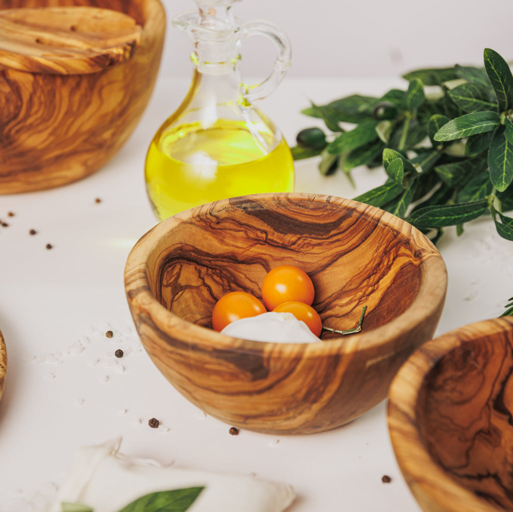 Small olive wood bowl holding yellow cherry tomatoes, styled with a bottle of olive oil, peppercorns, and olive branches on a white surface