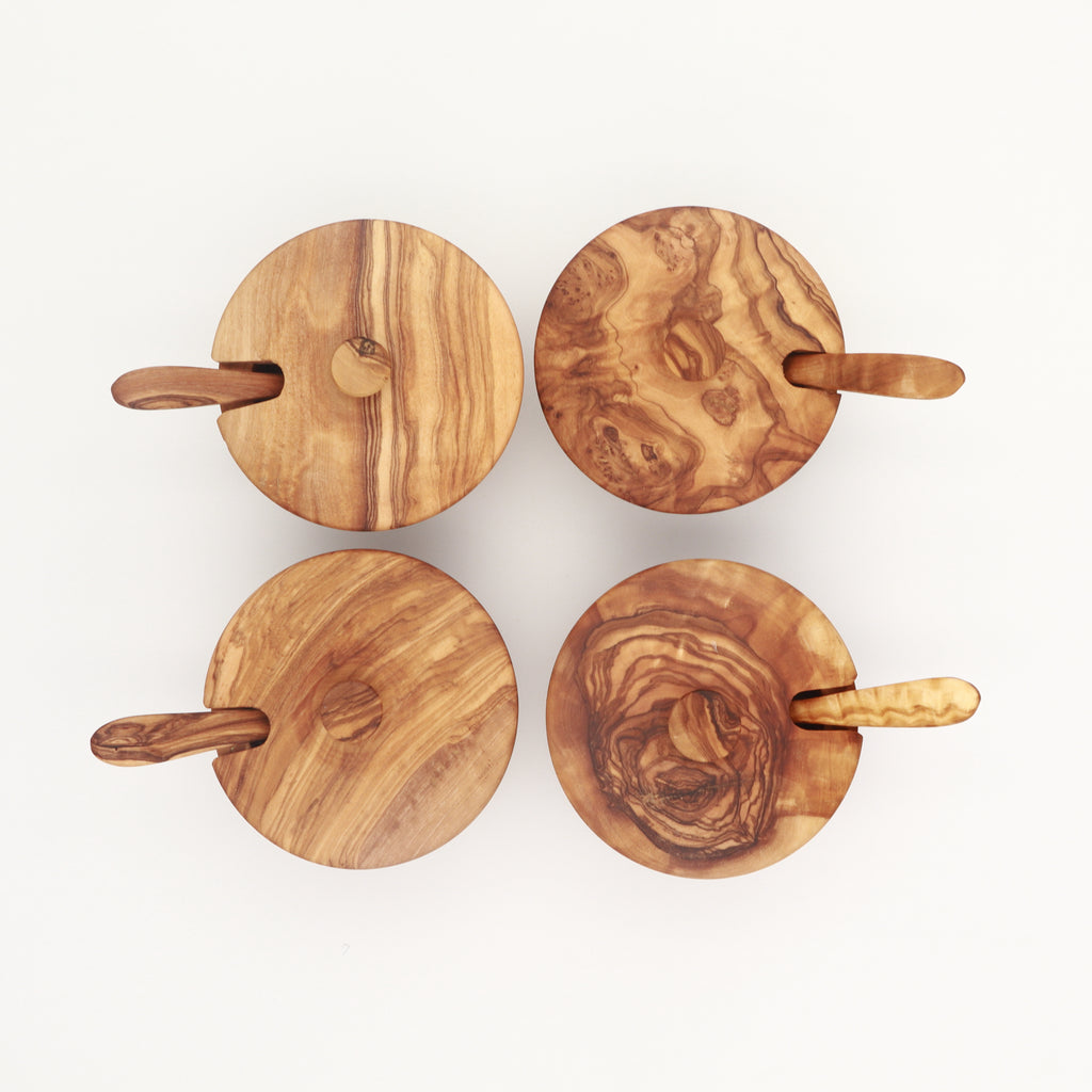 Four handcrafted olive wood spice boxes with swivel lids and small spoons, showing varied grain patterns.