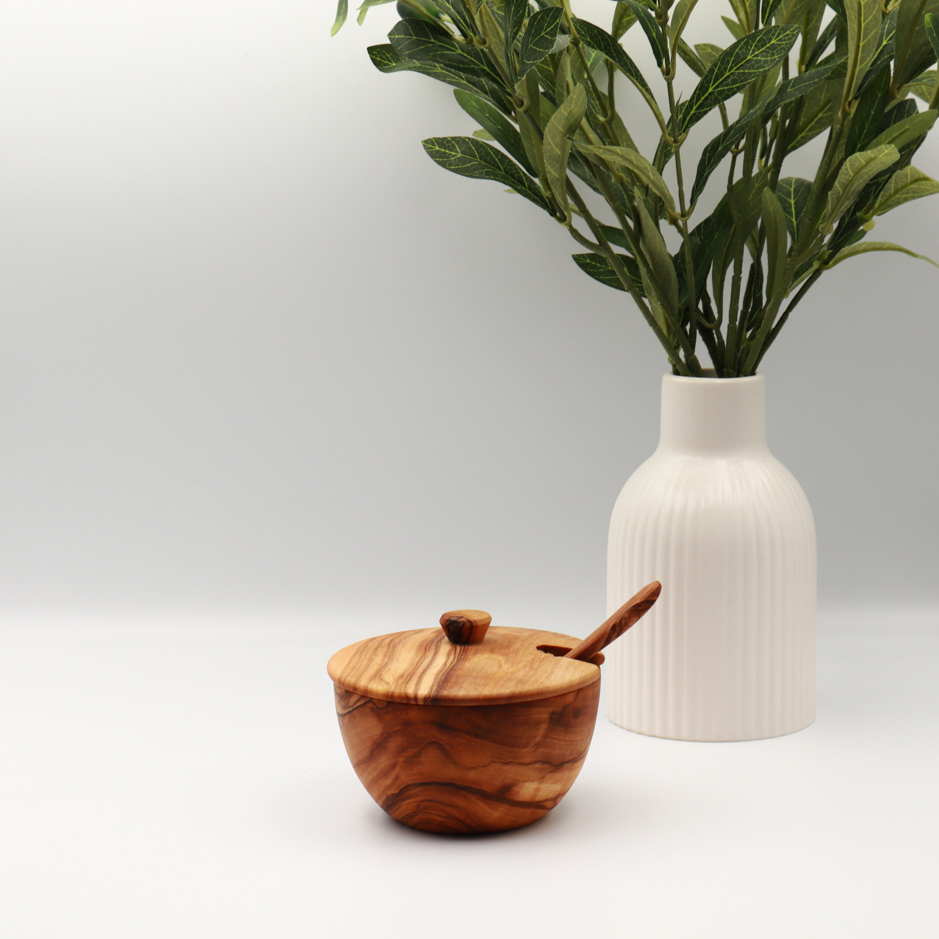 Single handcrafted Tunisian olive wood spice box with lid and serving spoon, styled on a white counter beside a vase of olive branches.
