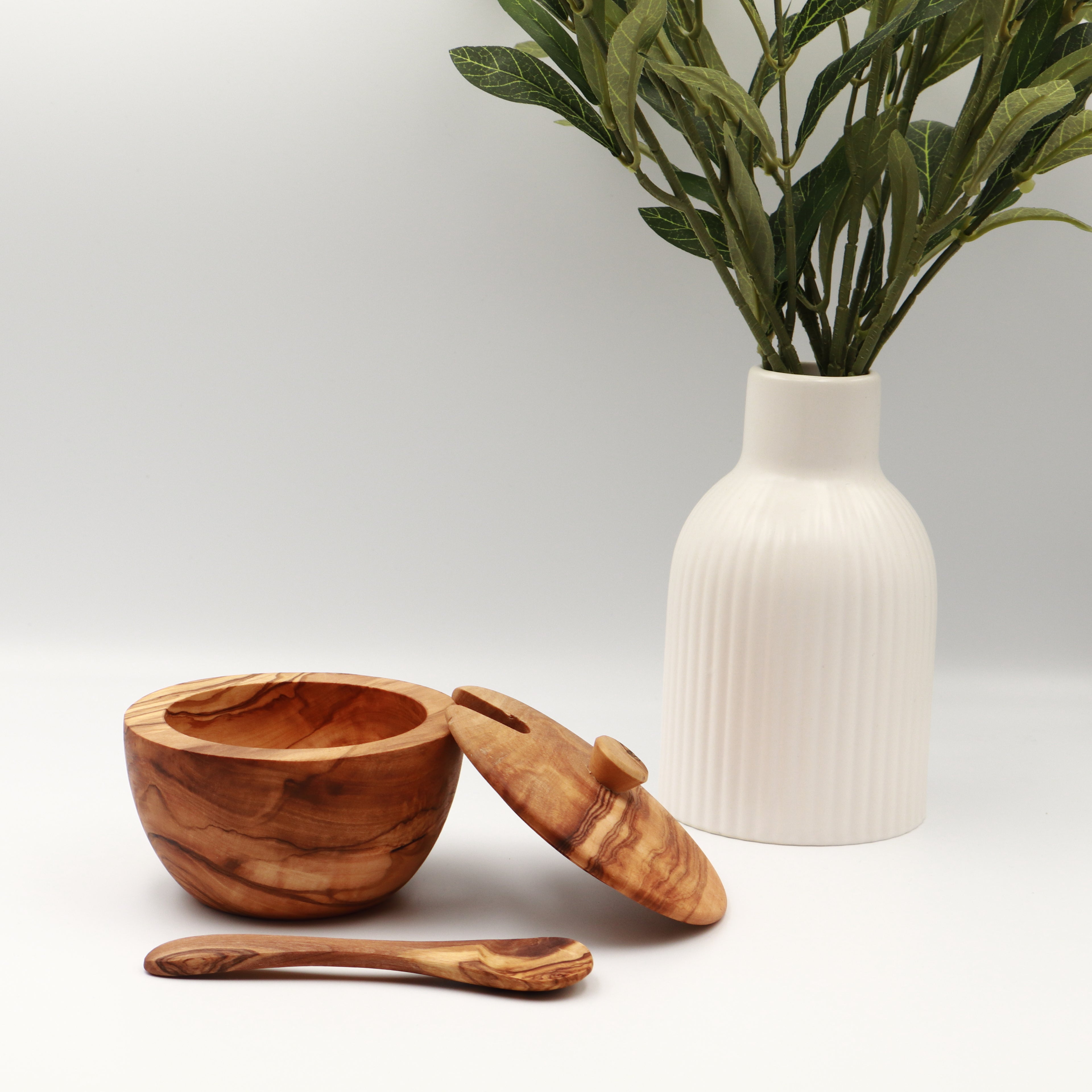 Single handcrafted Tunisian olive wood spice box with the lid removed and a serving spoon, styled on a white counter beside a vase of olive branches.