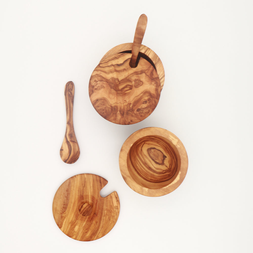 Overhead view of two handcrafted Tunisian olive wood spice boxes, one closed and one open with its lid and serving spoon beside it, on a white background.