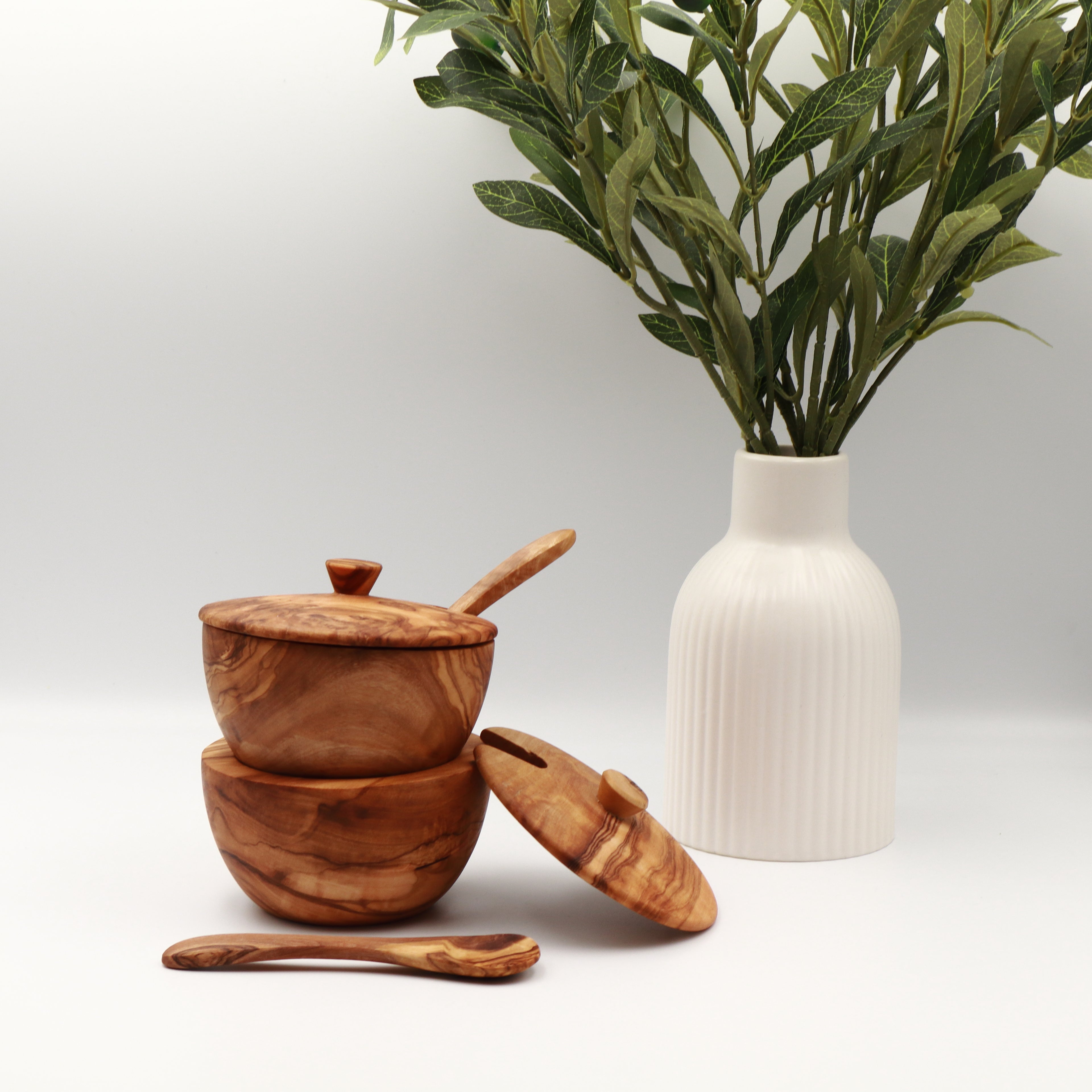 Two stacked handcrafted Tunisian olive wood spice boxes with lids and serving spoons, styled on a white counter beside a vase of olive branches.