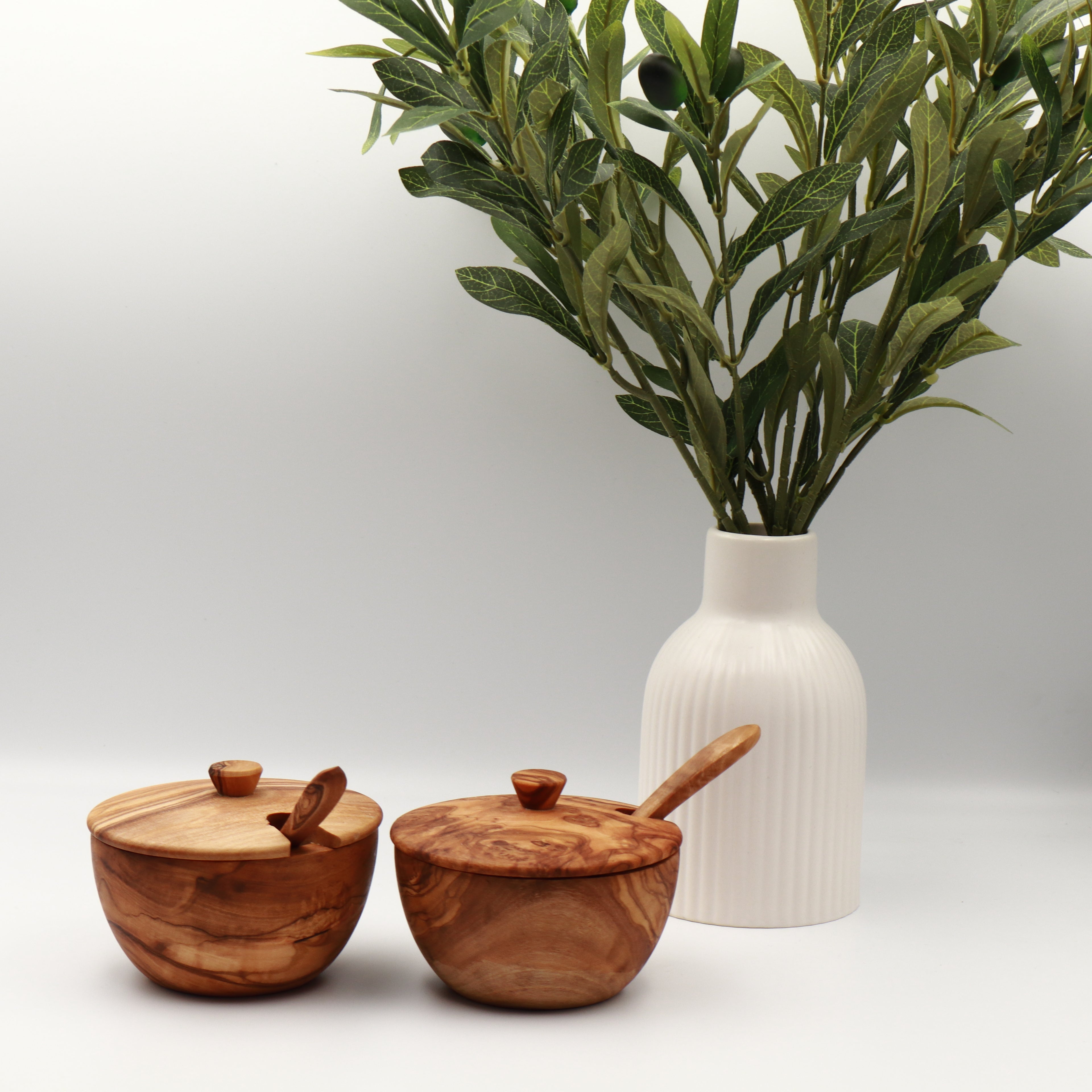 Two handcrafted Tunisian olive wood spice boxes with lids and serving spoons, styled on a white counter beside a vase of olive branches.