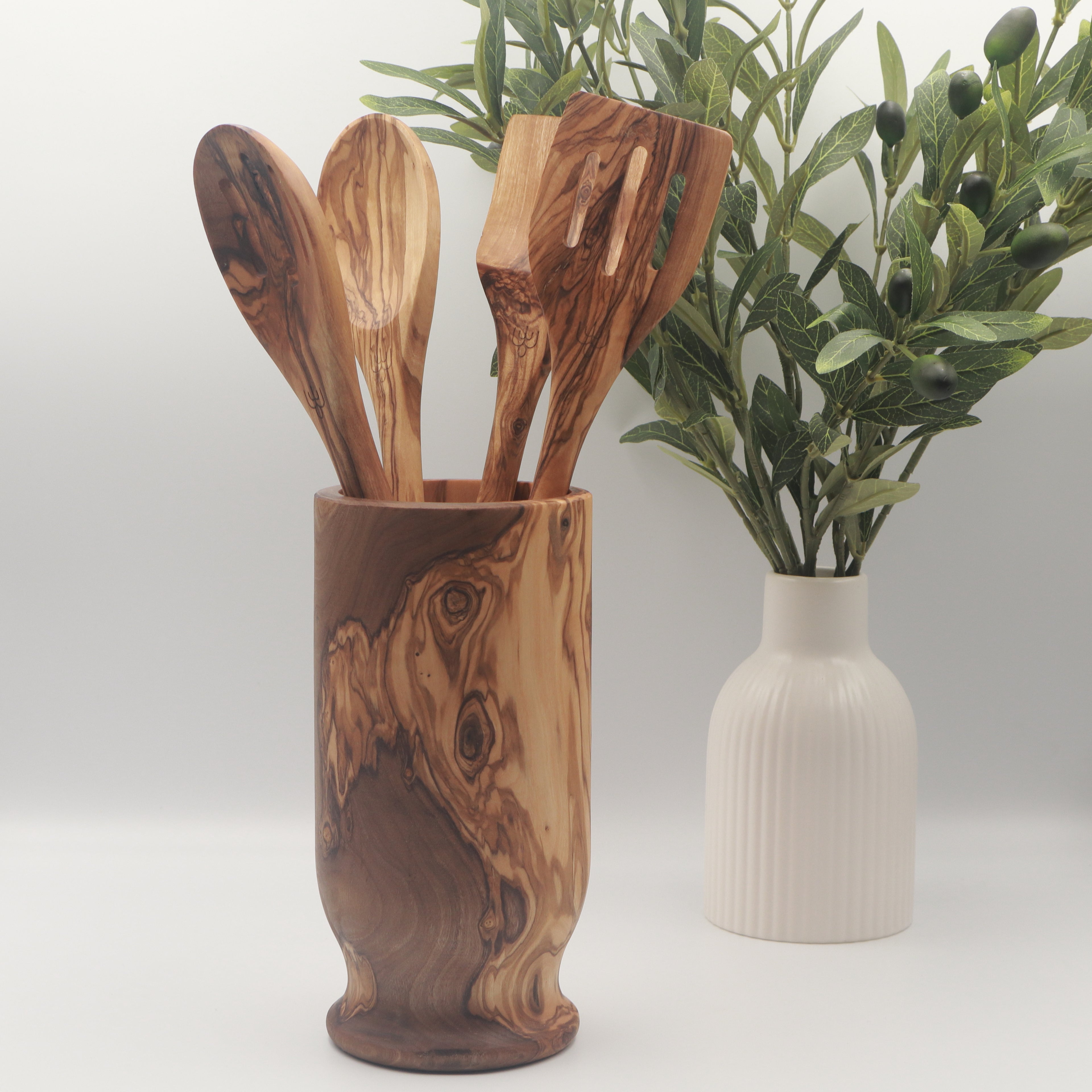 Handcrafted Tunisian olive wood utensil holder filled with four cooking utensils, styled on a white counter beside a vase of olive branches.