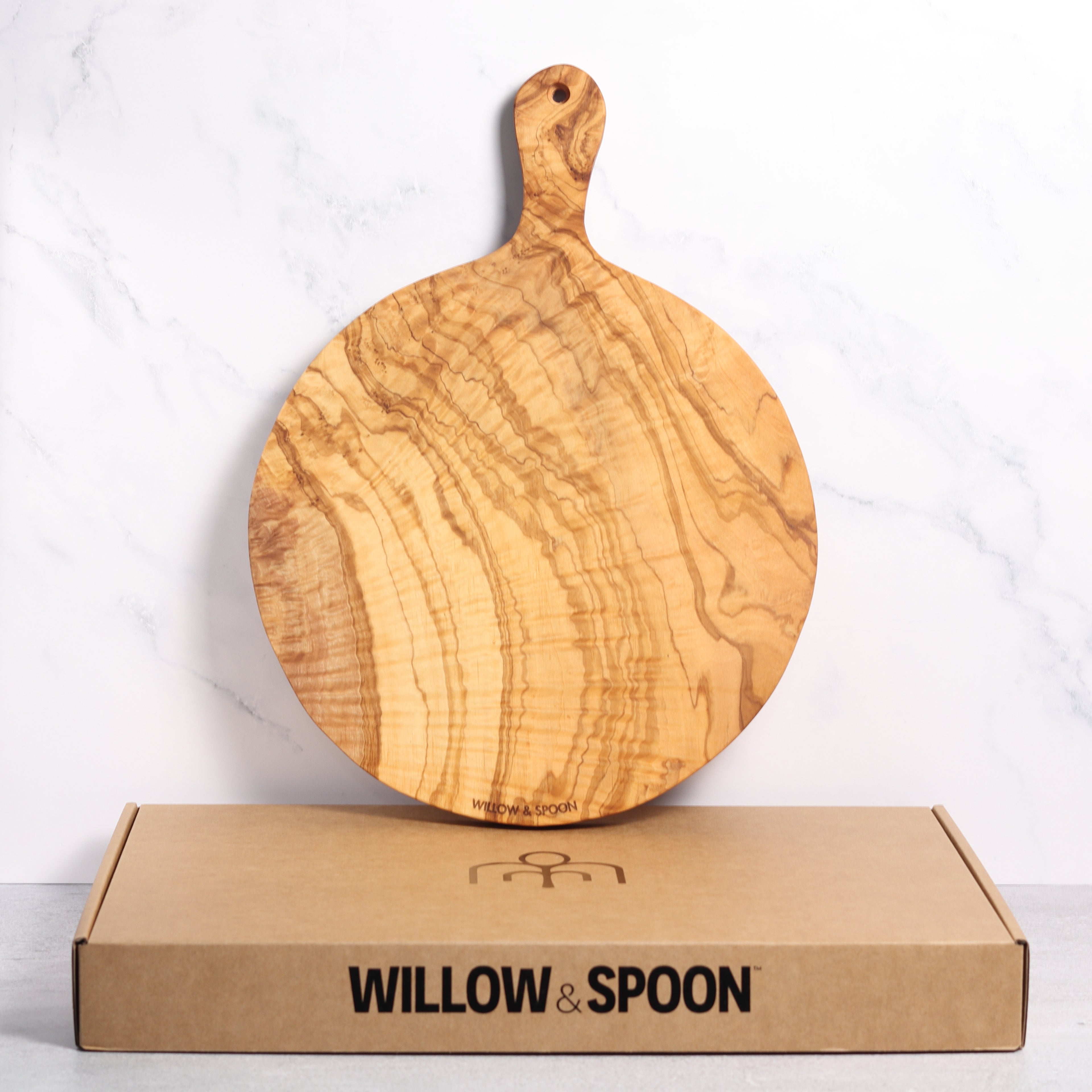 Round olive wood serving board with handle displayed upright on a Willow & Spoon branded box against a marble background