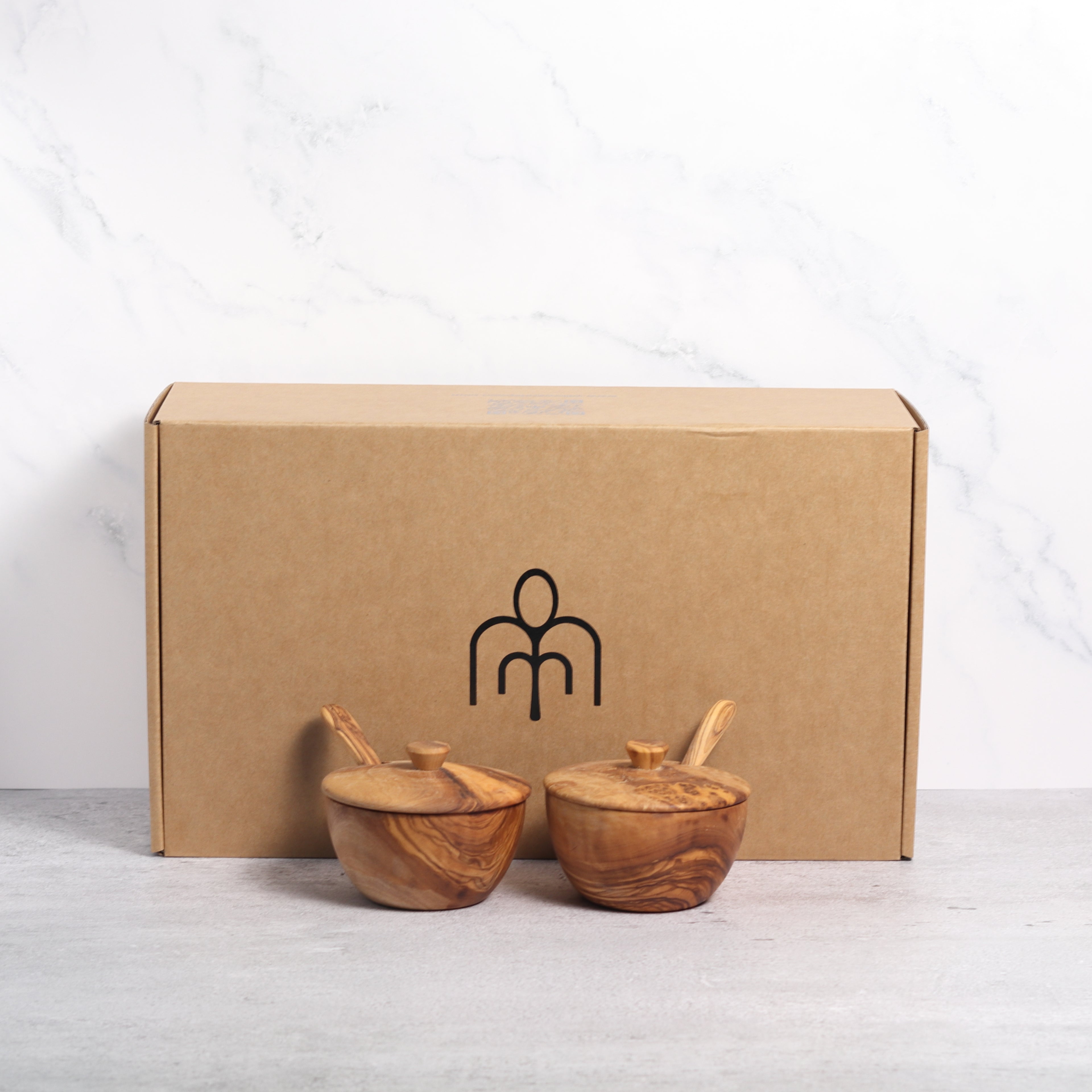 Two olive wood spice boxes with lids and spoons in front of a Willow & Spoon branded box against a marble background