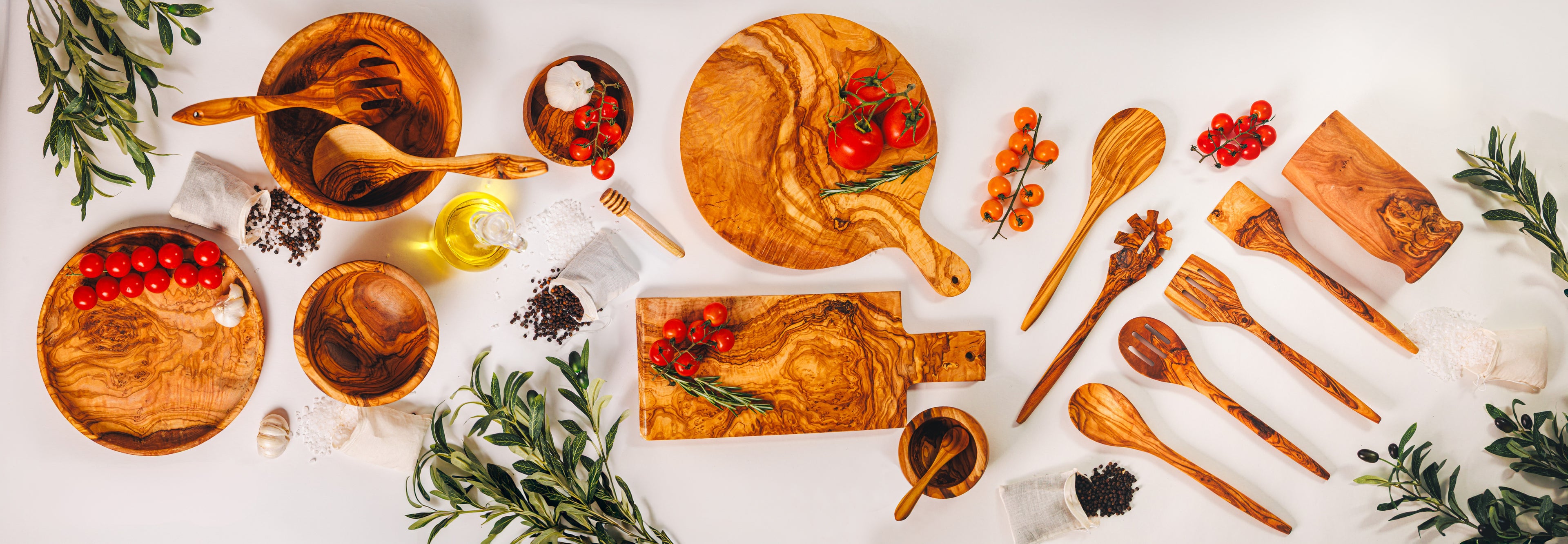 An overhead view of handcrafted olive wood kitchenware including round and rectangular cutting boards, nested bowls, and a variety of cooking utensils with fresh tomatoes and herbs.